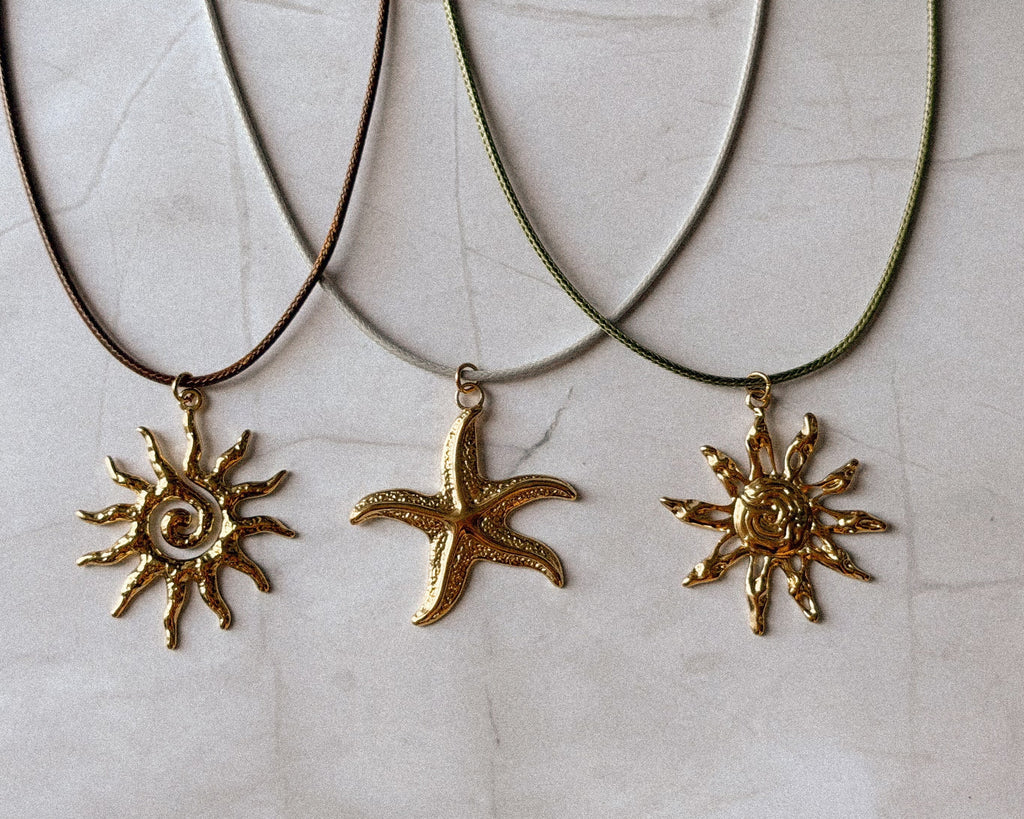 Three gold-toned jewelry pendants laid on a light beige textured surface: two sunburst charms and one starfish charm, each attached to a cord necklace.
