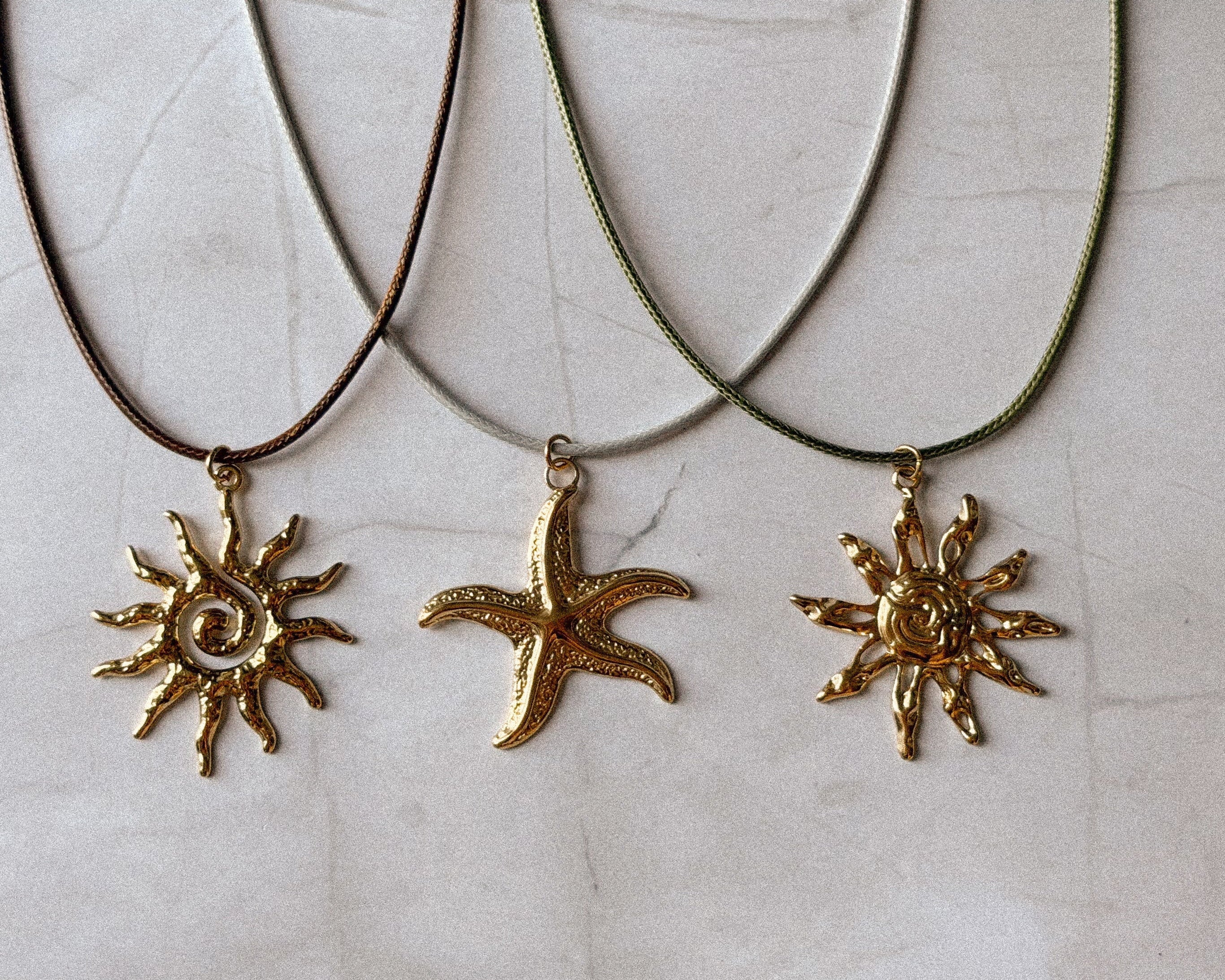 Three gold-toned jewelry pendants laid on a light beige textured surface: two sunburst charms and one starfish charm, each attached to a cord necklace.