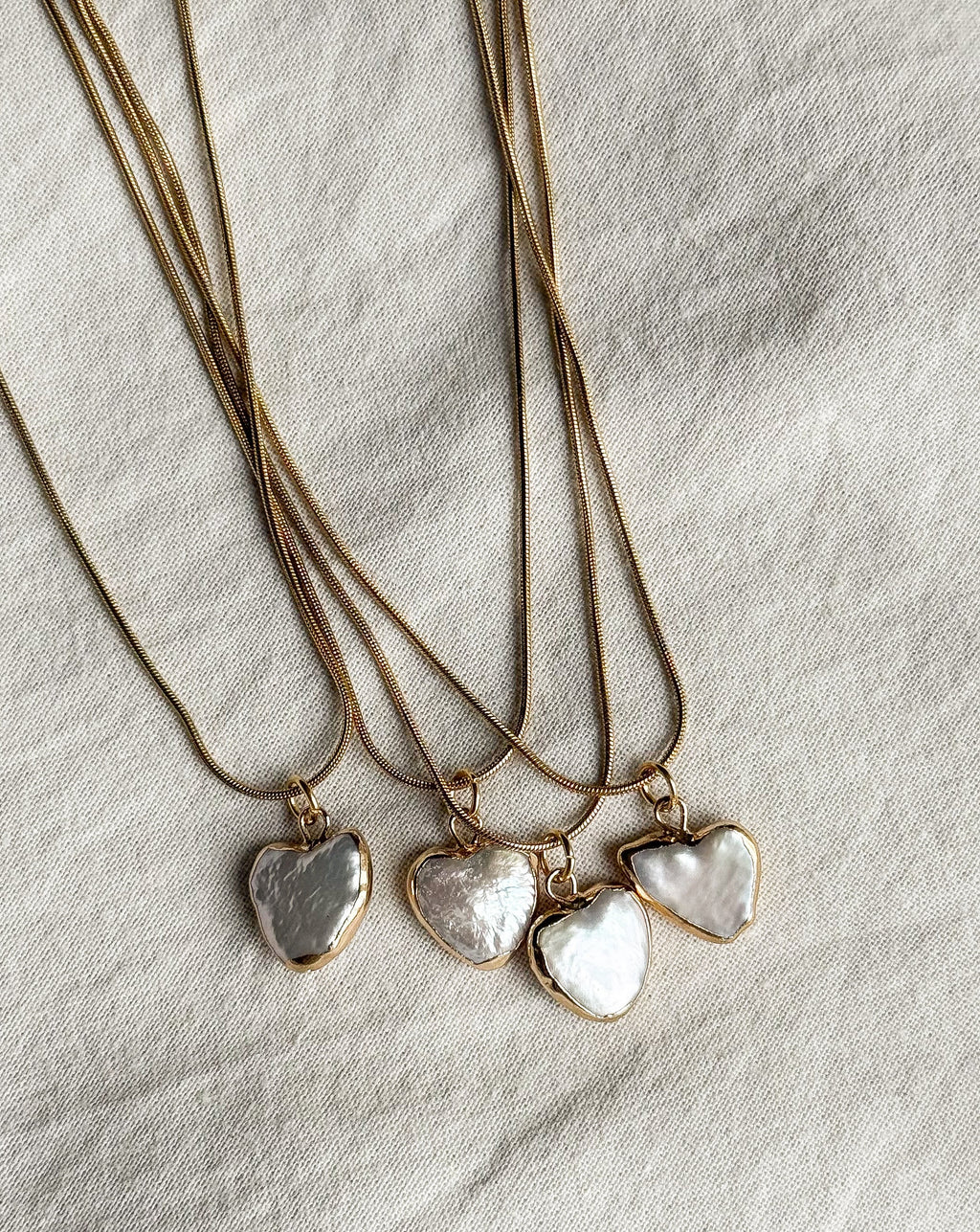 Three gold necklaces with heart-shaped pearl pendants on a neutral fabric background