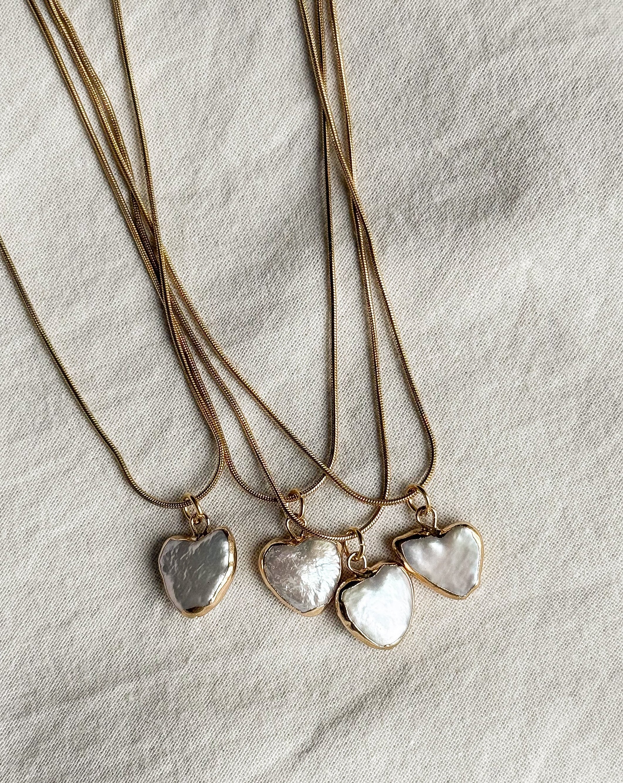 Three gold necklaces with heart-shaped pearl pendants on a neutral fabric background